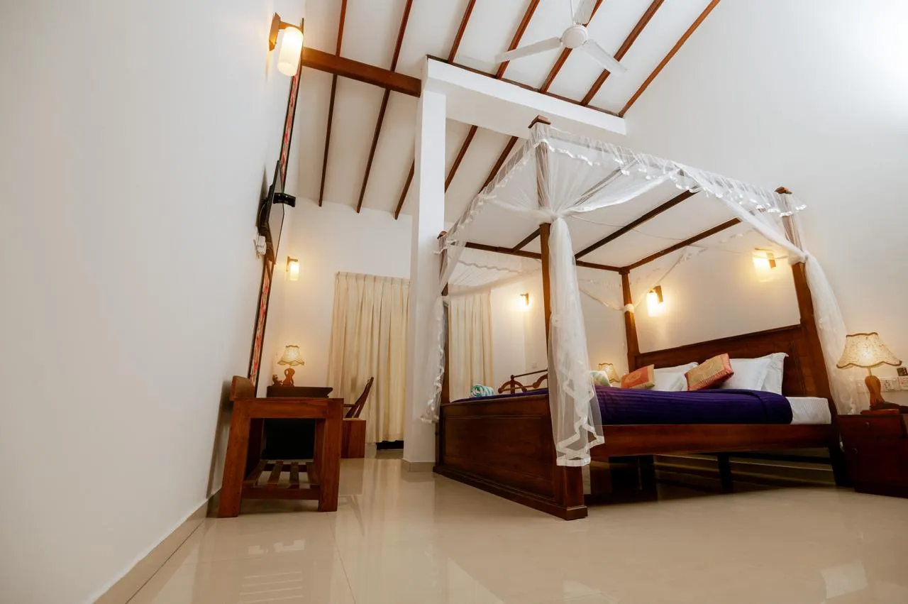 A four-poster bed and wooden desk beneath a high ceiling with exposed beams